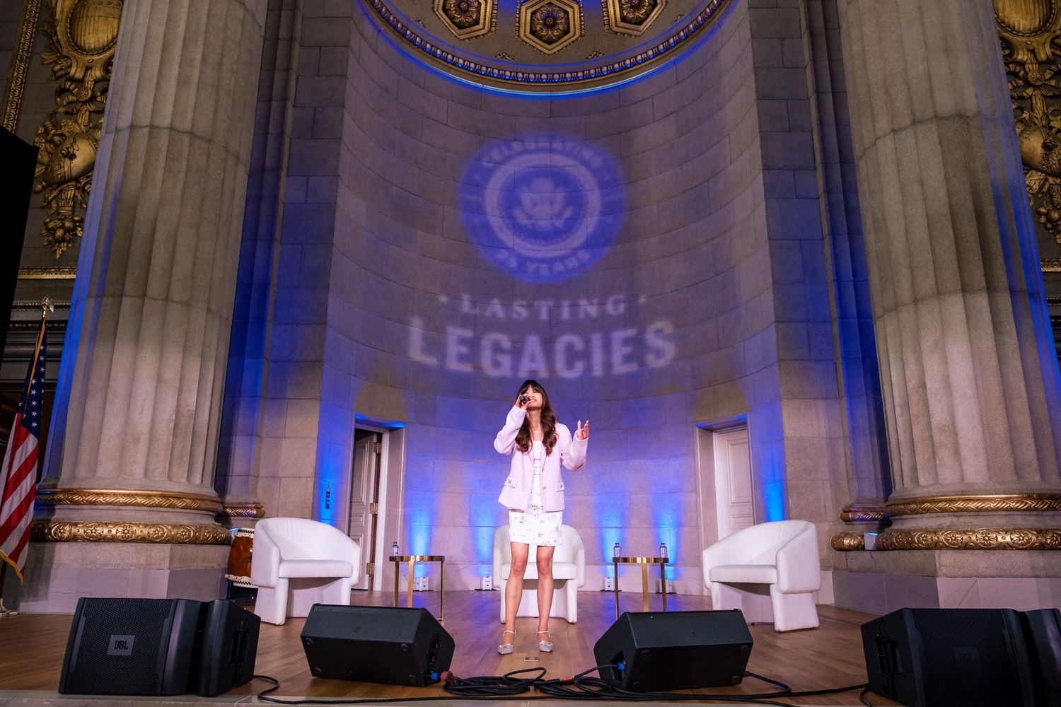 Woman in white outfit performs on stage at Lasting Legacies event beneath grand columns.