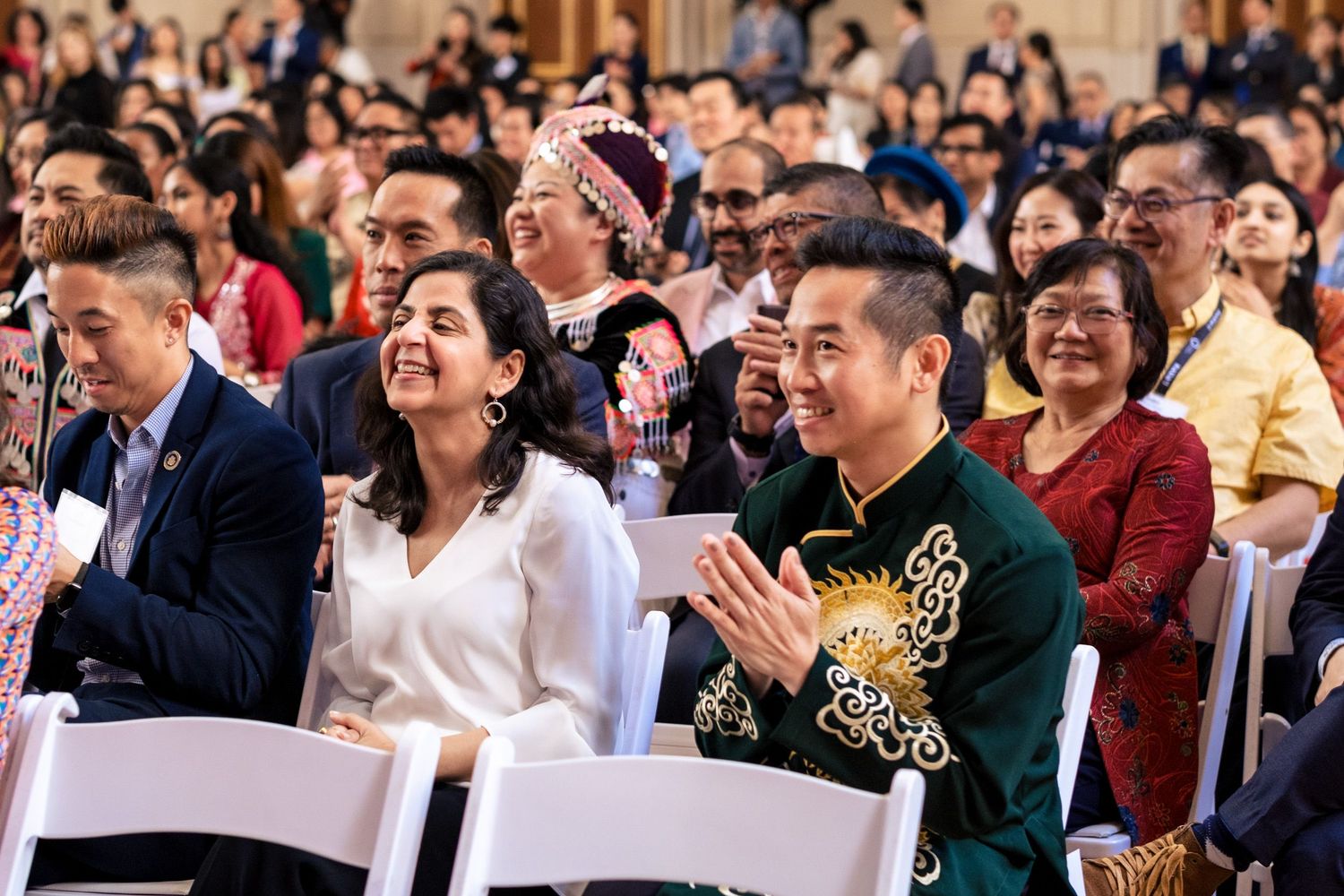 Diverse group of smiling people in traditional and formal attire applauding at a multicultural celebration event.