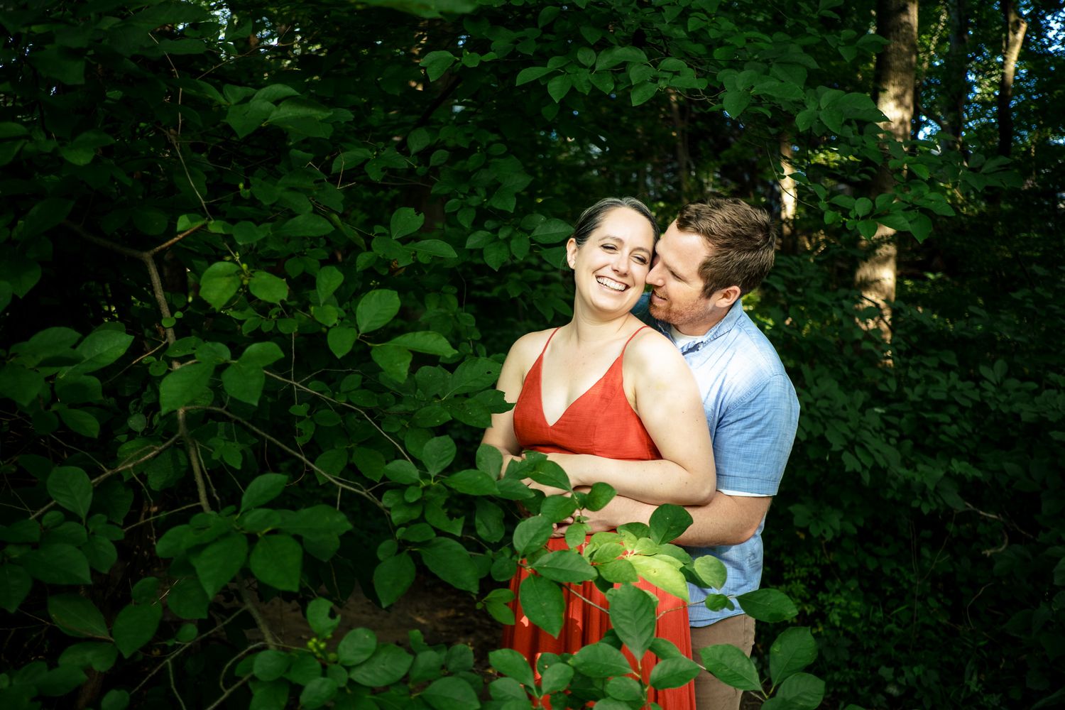 Couple embracing and smiling together surrounded by lush green foliage in a wooded area.