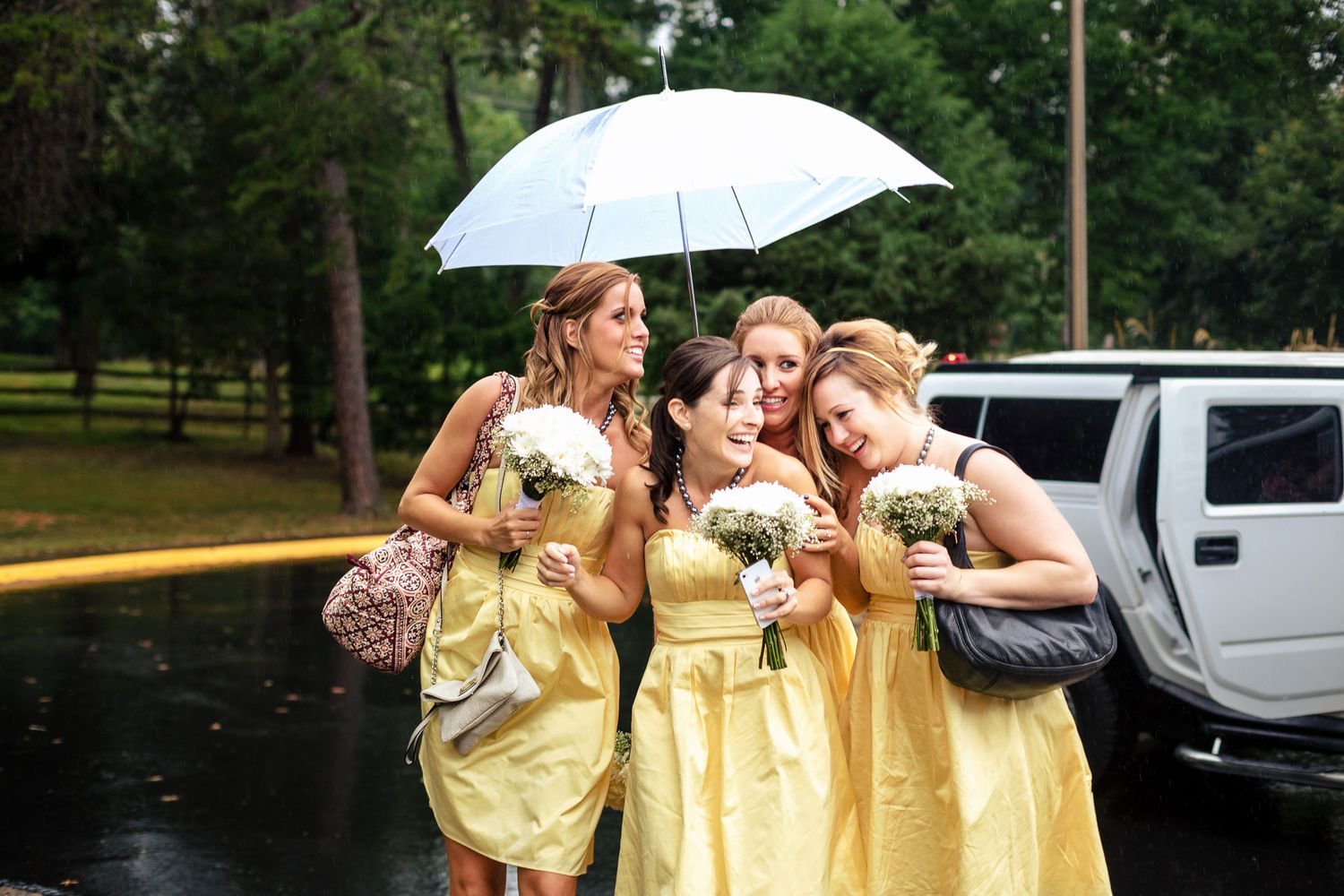 Four bridesmaids in yellow dresses huddle under an umbrella in the rain.