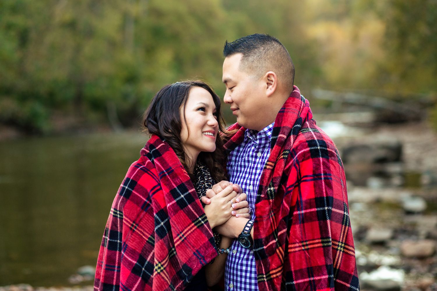 Couple wrapped together in a red plaid blanket, smiling at each other near a creek.