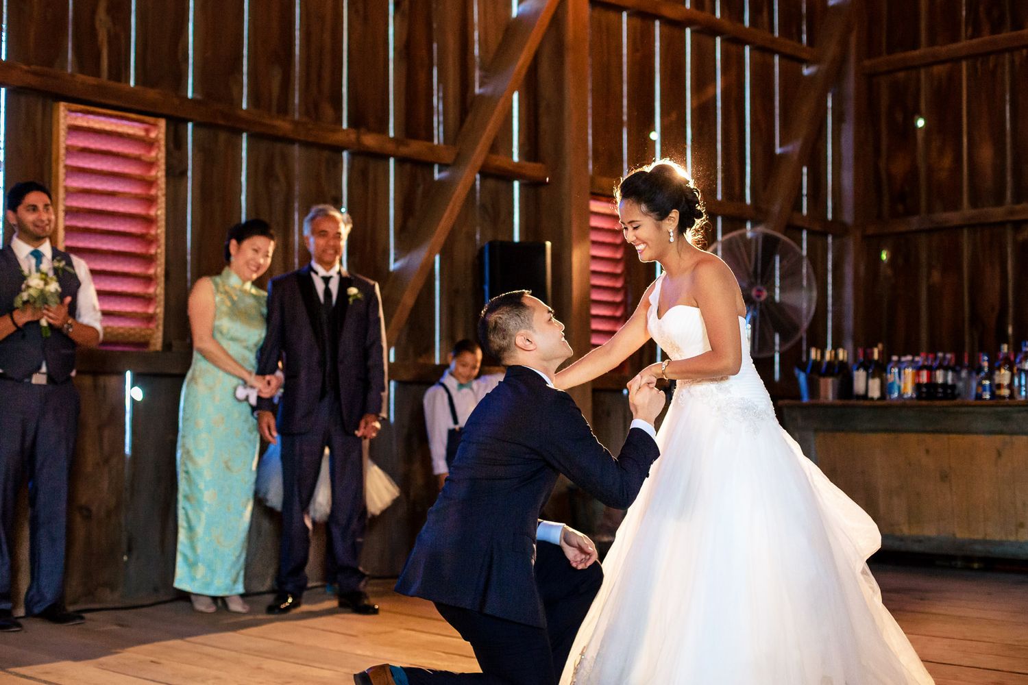 Groom kneels dramatically before his bride during their first dance in a warmly lit rustic barn.