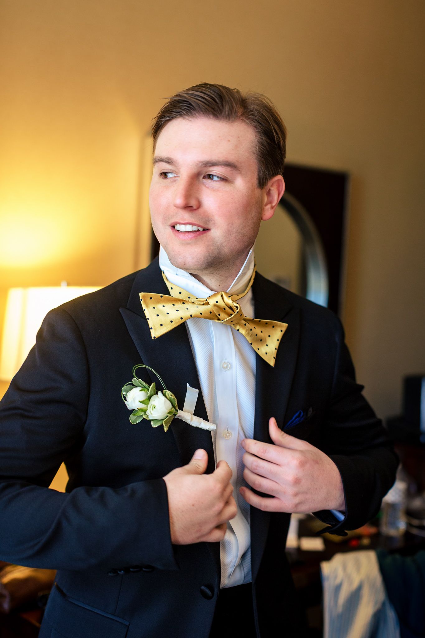 Groom in black tuxedo with gold bow tie and boutonniere adjusting his jacket indoors.