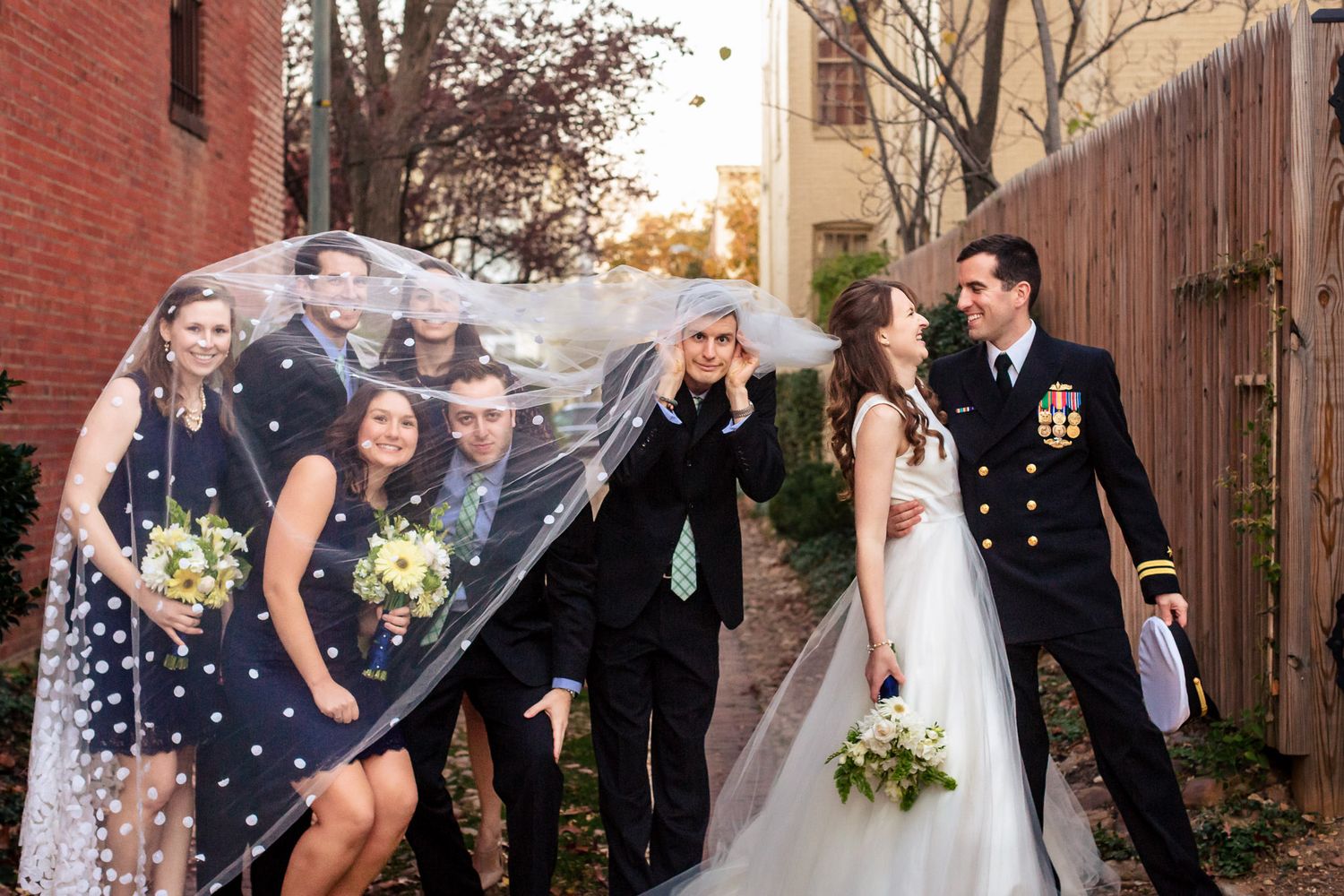 Wedding party laughs as bride's veil blows in the wind outdoors.