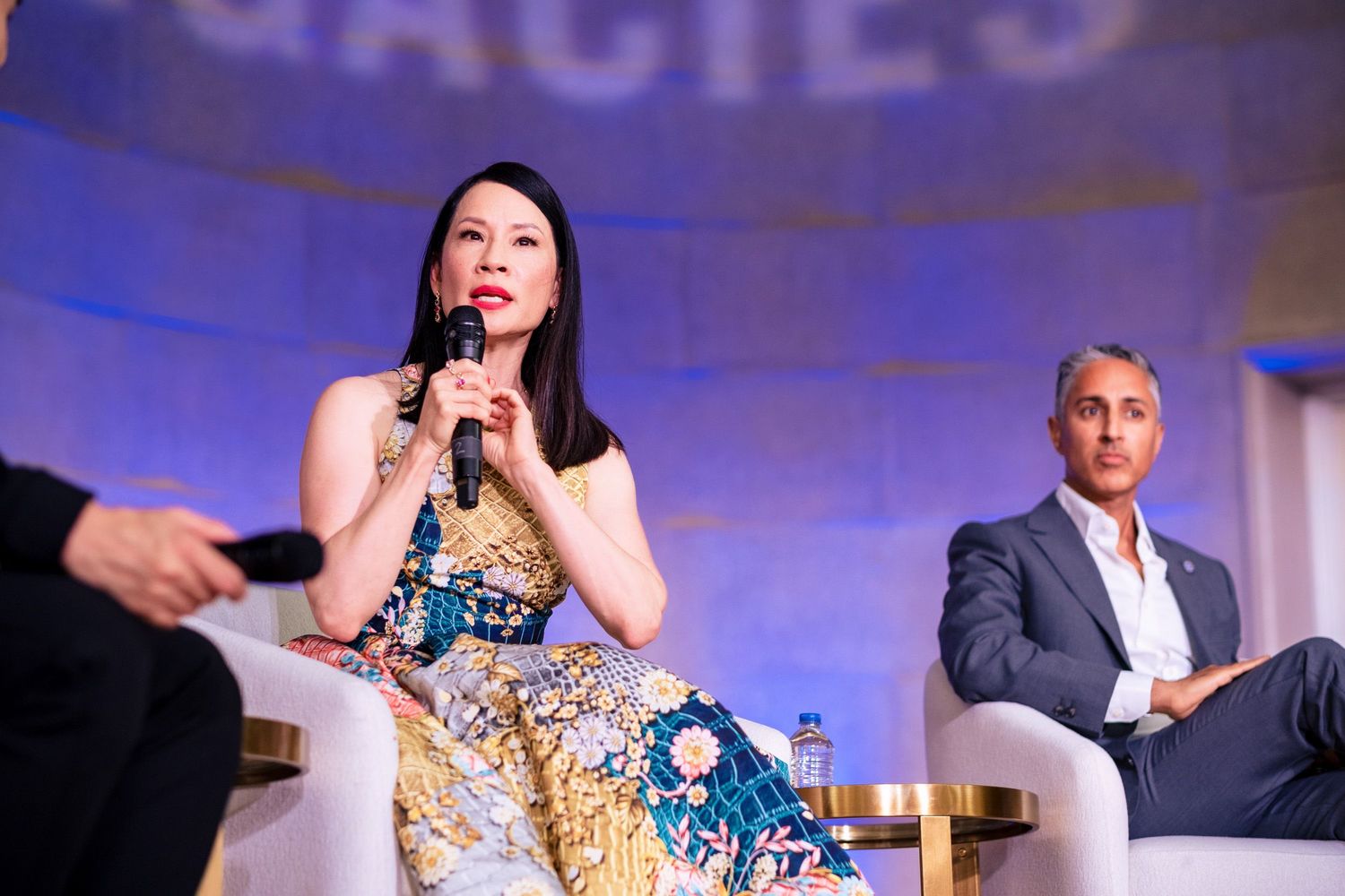 Woman in floral dress speaks at panel event, man in gray suit seated behind her on stage.
