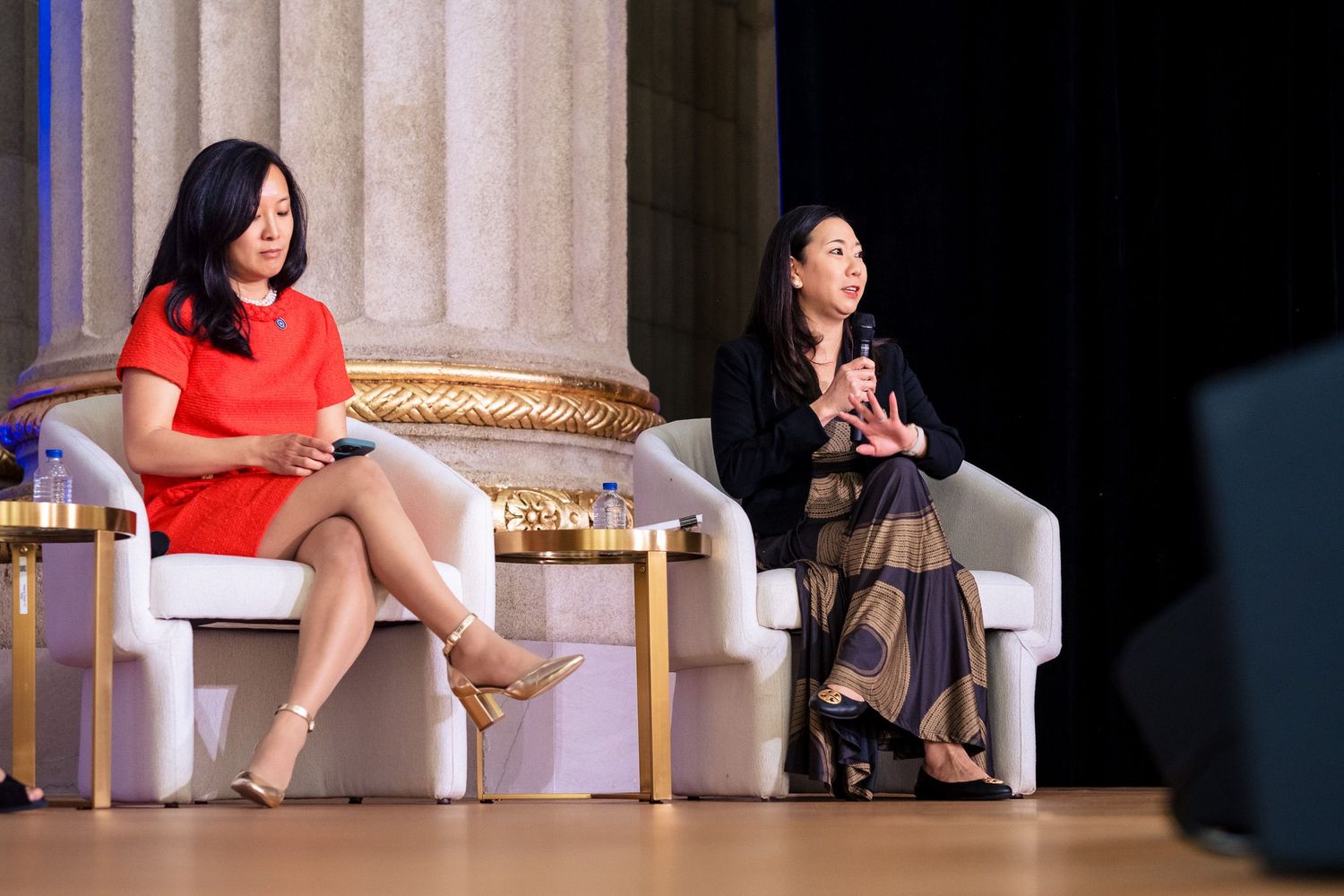 Two women seated on white chairs on stage during a panel discussion at a formal event.