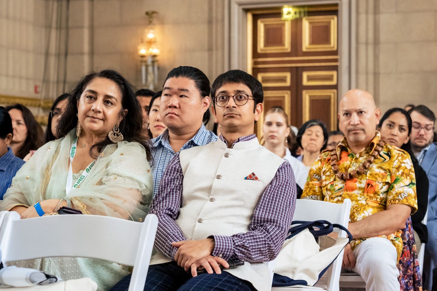 Diverse audience seated in rows at an indoor event, attentively watching a presentation in a grand hall.