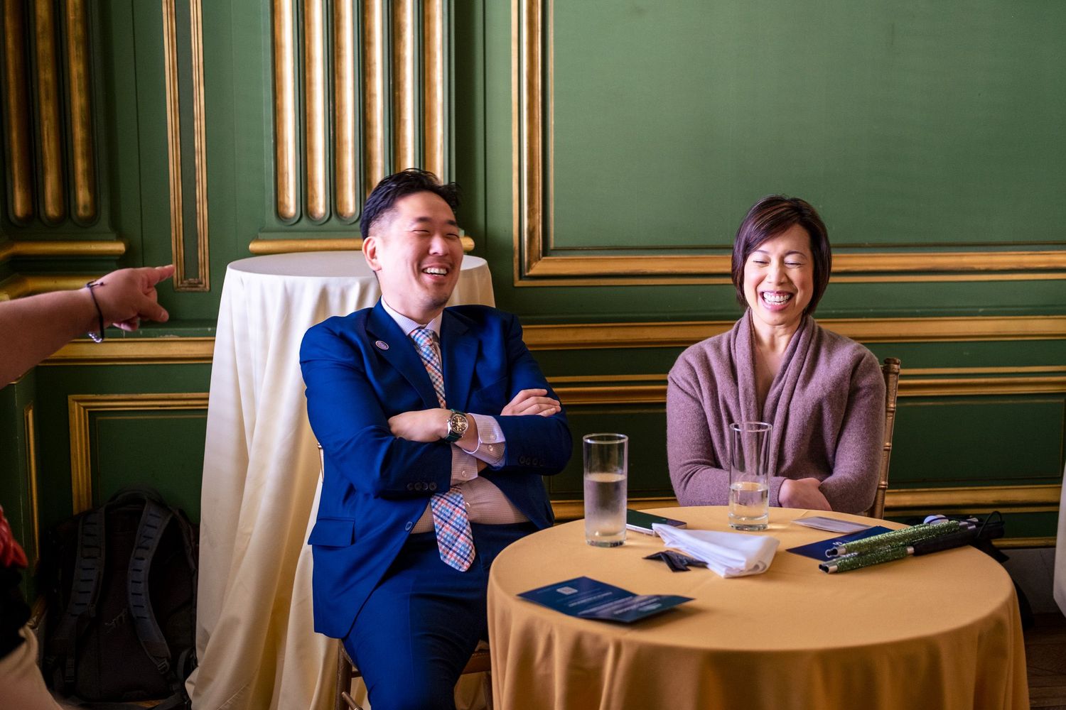 Two people smiling and laughing at a round table in an elegant green and gold room during a formal event.