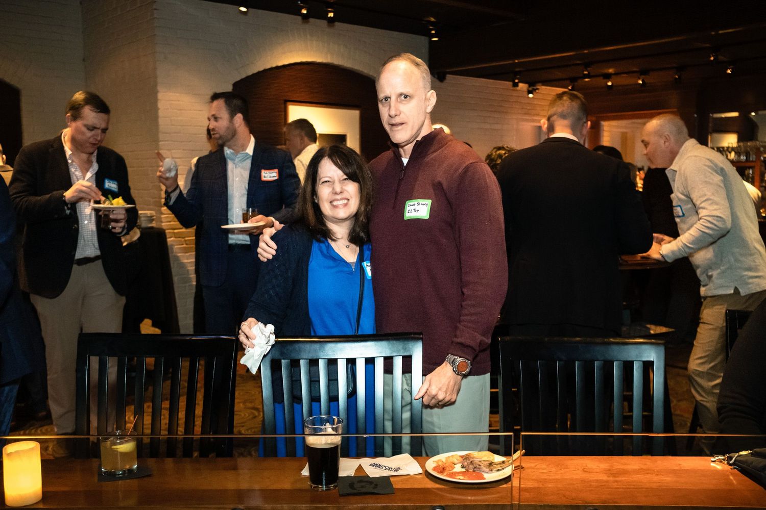 A smiling couple poses together at a casual networking event in a modern bar setting with guests mingling in the background.