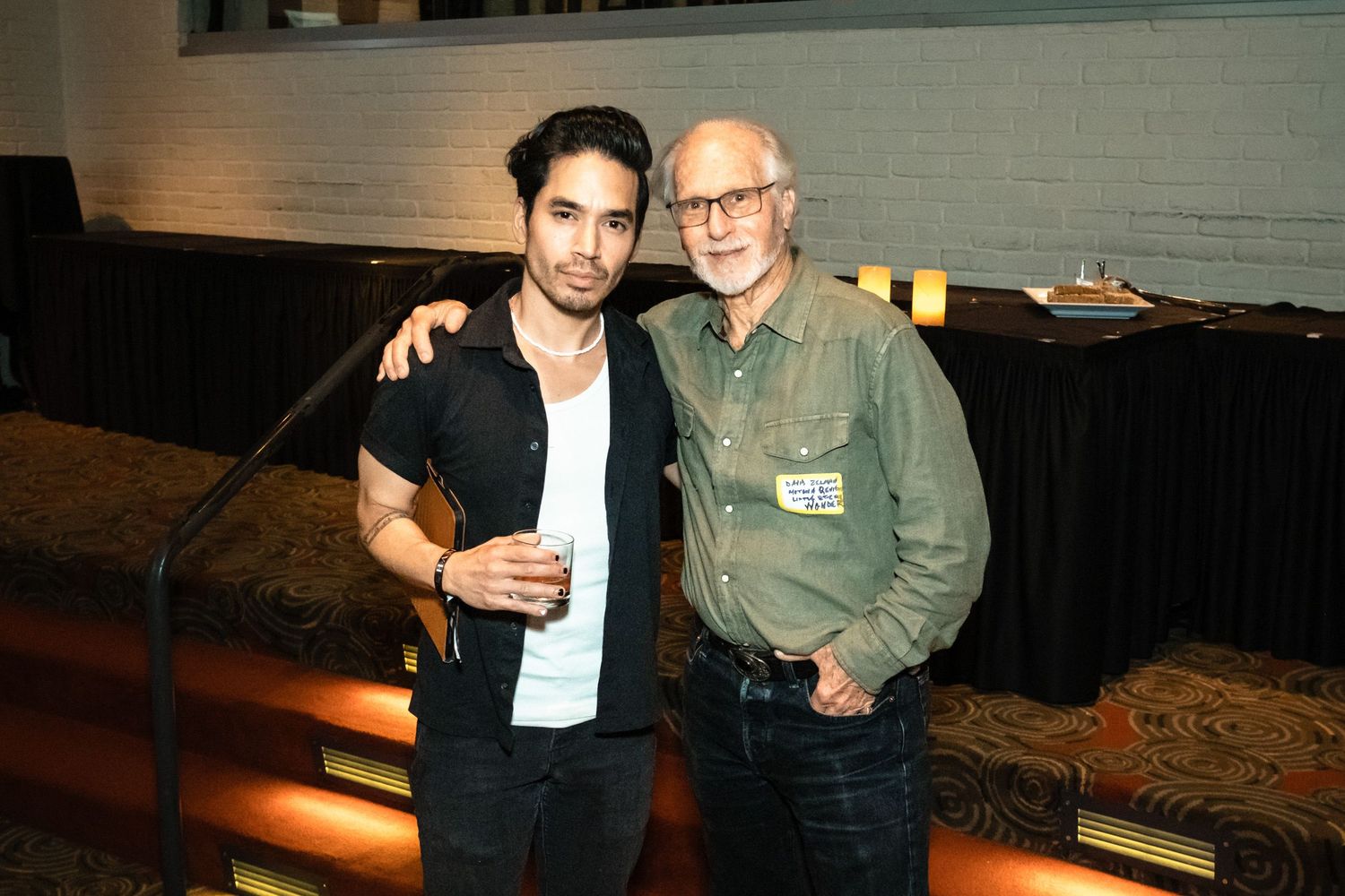 Two men pose together smiling at an indoor event, one holding a drink, against a dark auditorium background.