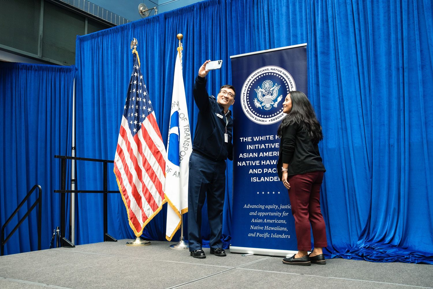 Two people take a selfie on stage near US and government flags at a White House AANHPI Initiative event.
