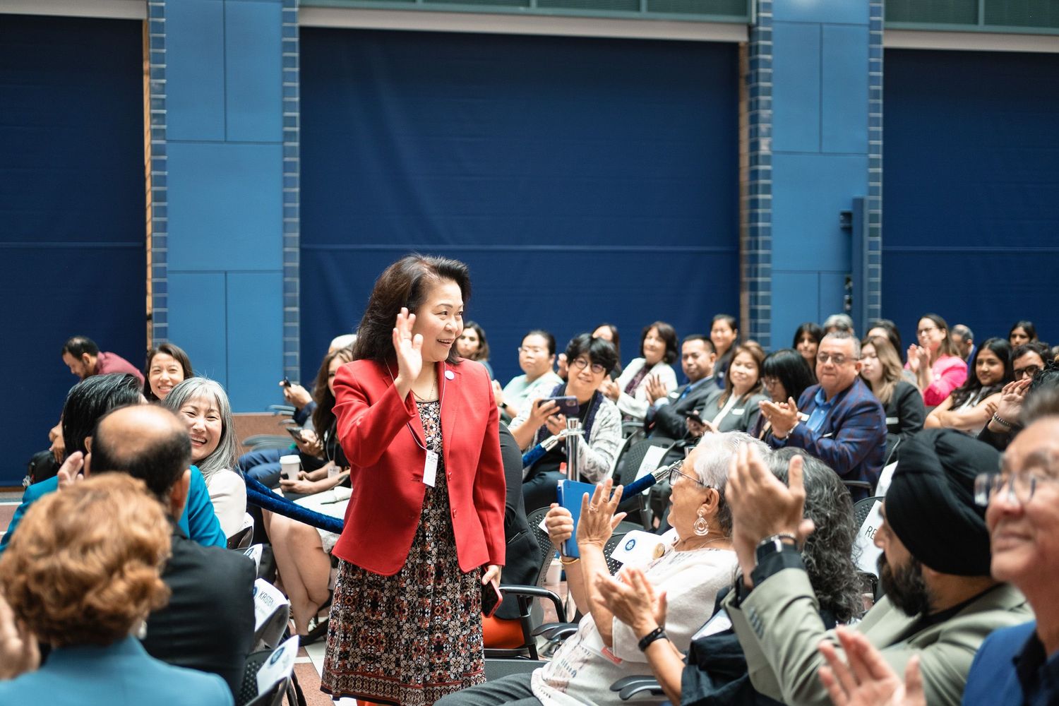A woman in a red blazer speaks on phone at a crowded indoor event with diverse audience members seated and applauding.