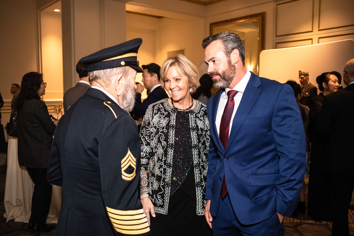 Three elegantly dressed guests converse at a formal event, including a man in military uniform and a woman in a patterned jacket.