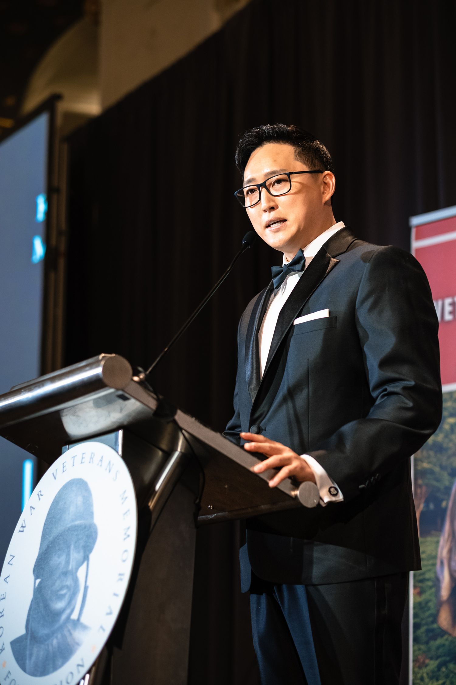 A man in a dark tuxedo with bow tie speaks at a podium during a formal veterans foundation gala event.