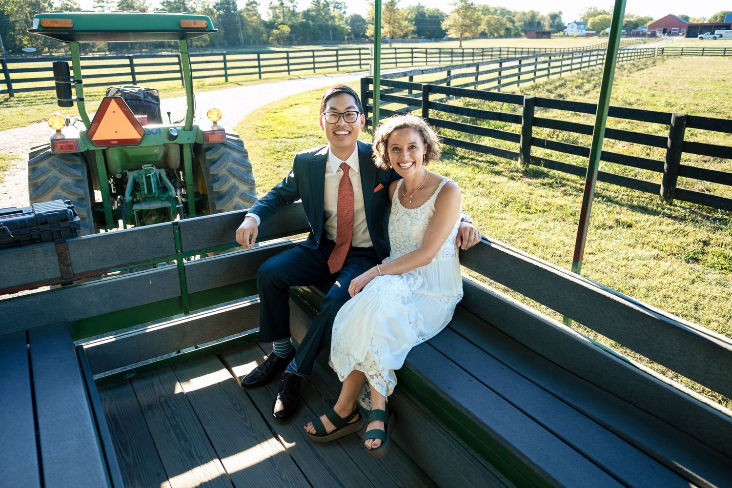 Bride and groom sitting together on a farm wagon with a green tractor and fenced pasture in the background.