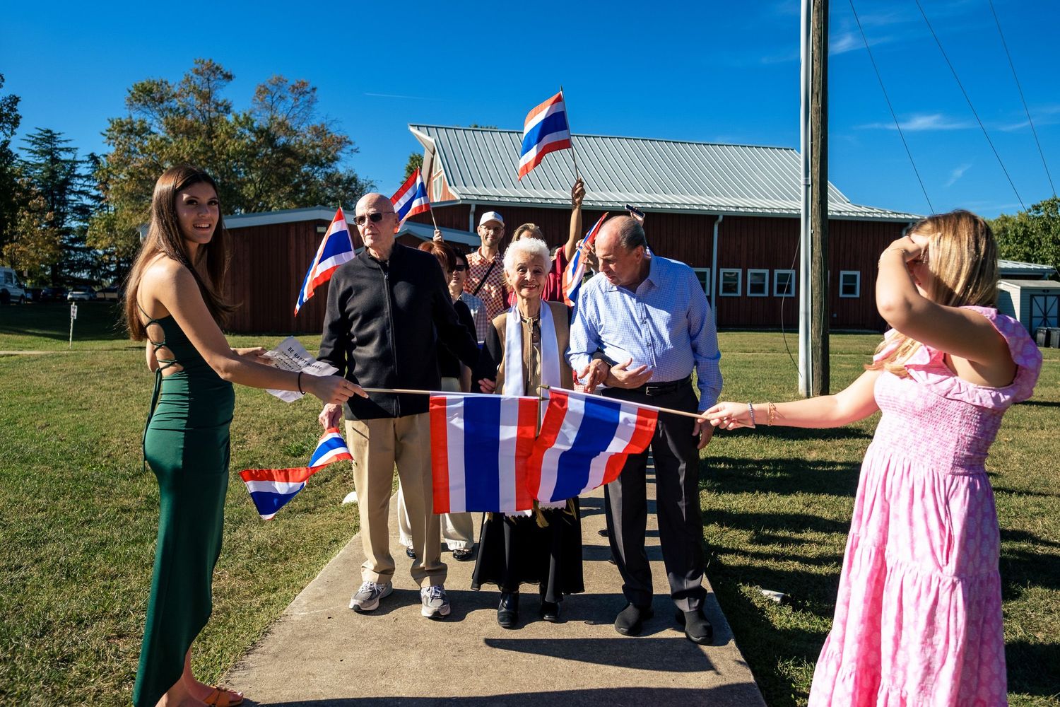 A group of smiling guests wave Thai and other flags during a colorful outdoor wedding celebration.