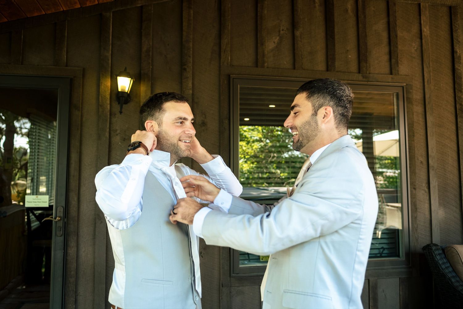 Two groomsmen in light gray suits helping each other with ties outside a wooden building.
