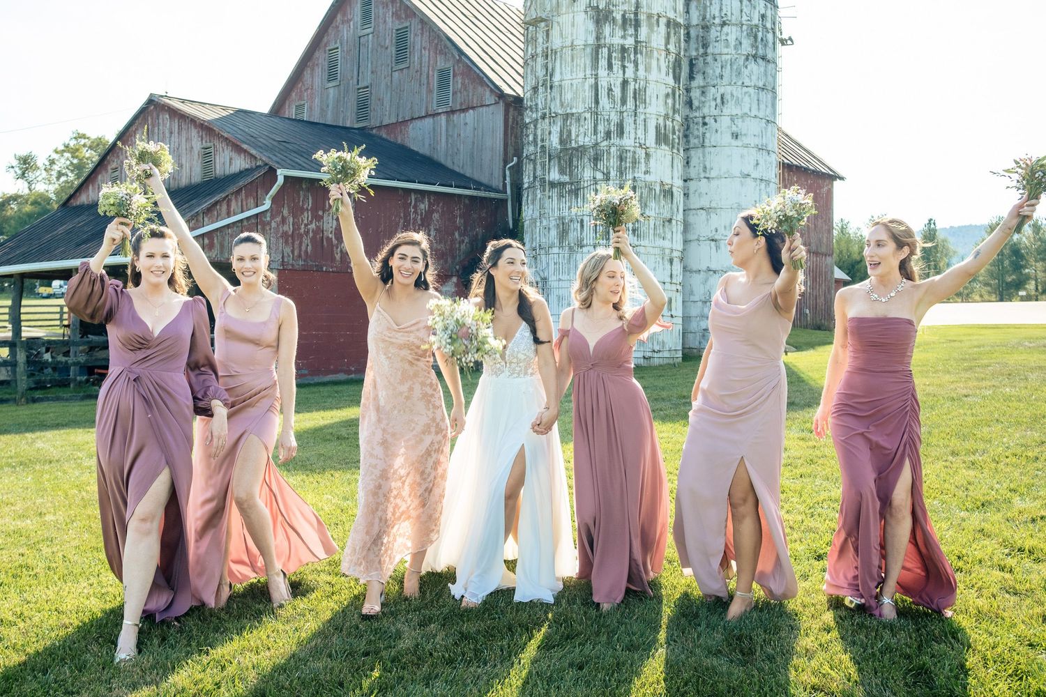 Bridesmaids in pink and mauve gowns celebrate with bride in white on a farm lawn near a red barn.
