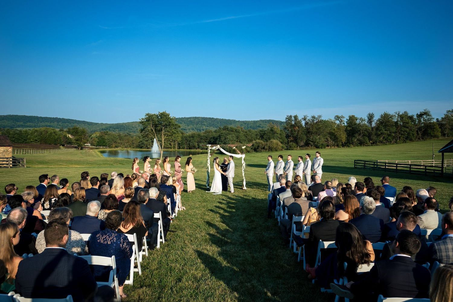 Aerial view of an outdoor wedding ceremony with guests seated in white chairs and a scenic mountain backdrop.