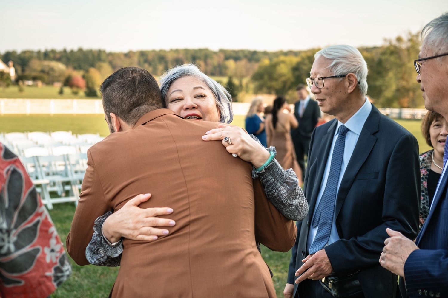 Groom in brown suit receives warm hug from an older woman guest following outdoor wedding ceremony.
