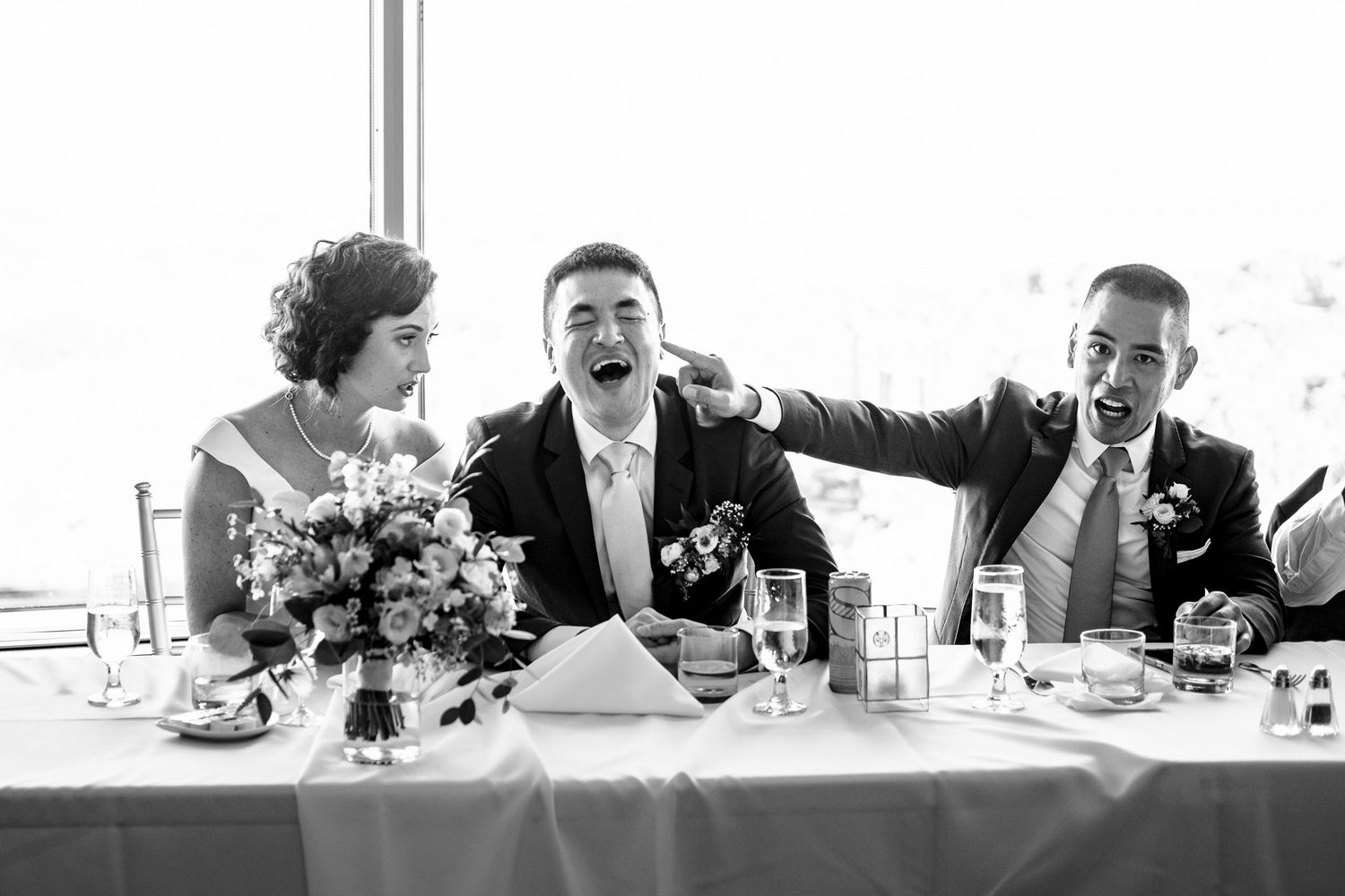 Three guests laughing joyfully together at elegant wedding reception dinner table.