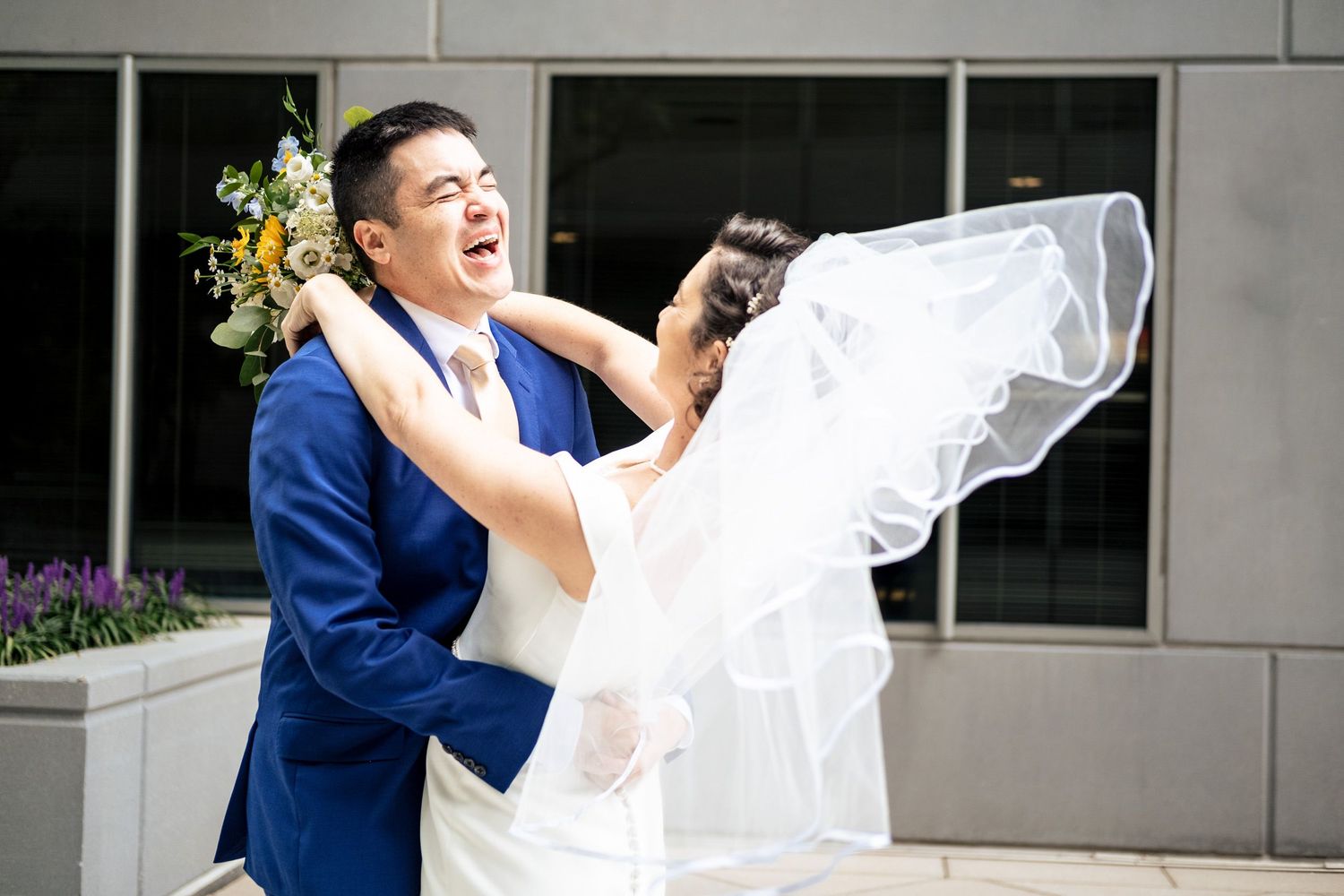 Groom in blue suit lifts bride in white wedding gown in an joyful embrace with veil flowing.