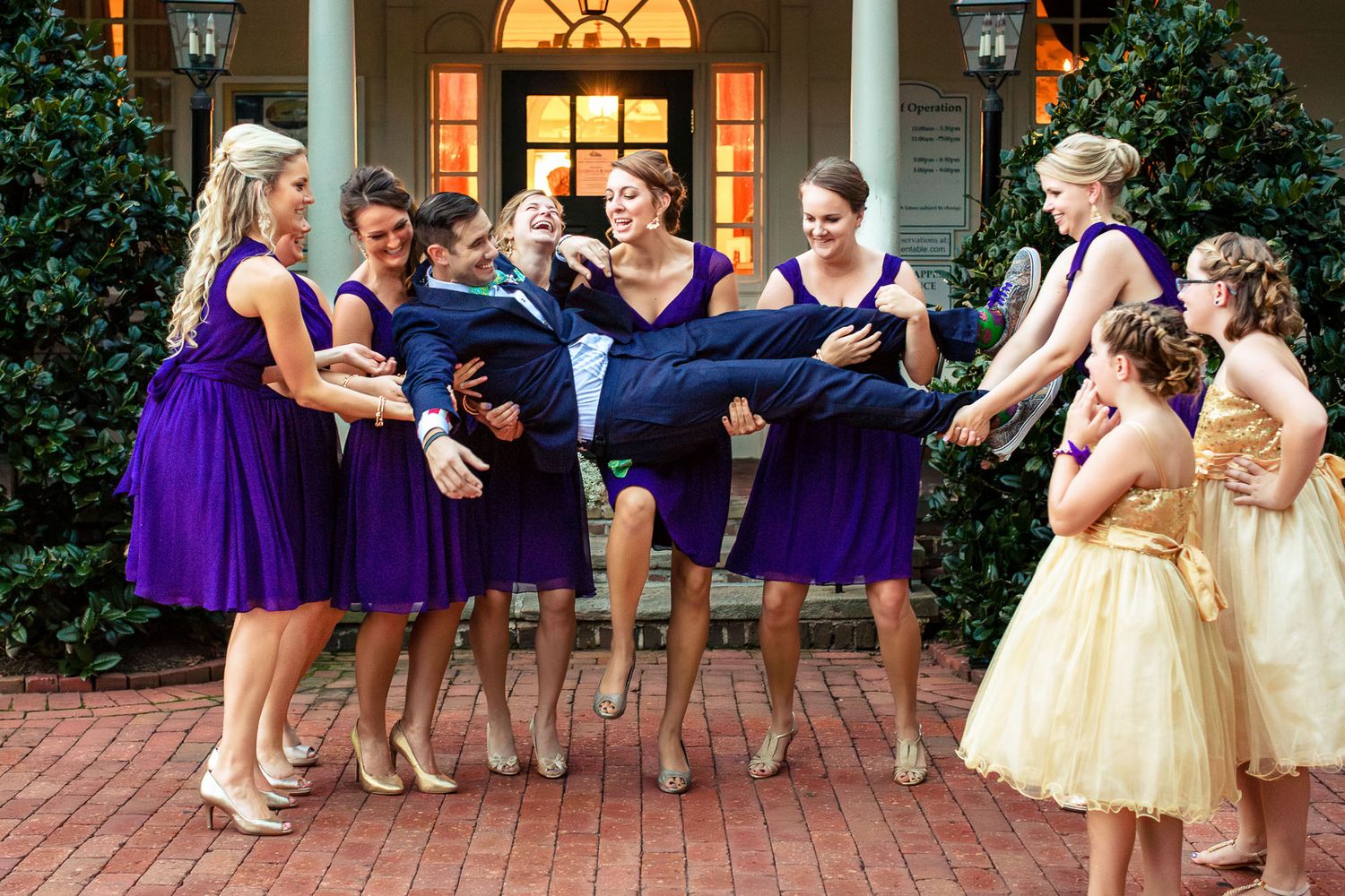 Laughing bridesmaids in purple dresses playfully carry the groom outside a elegant venue.