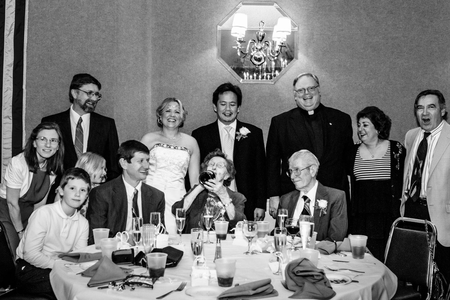 Black and white photo of wedding reception group gathered around a table with a priest.