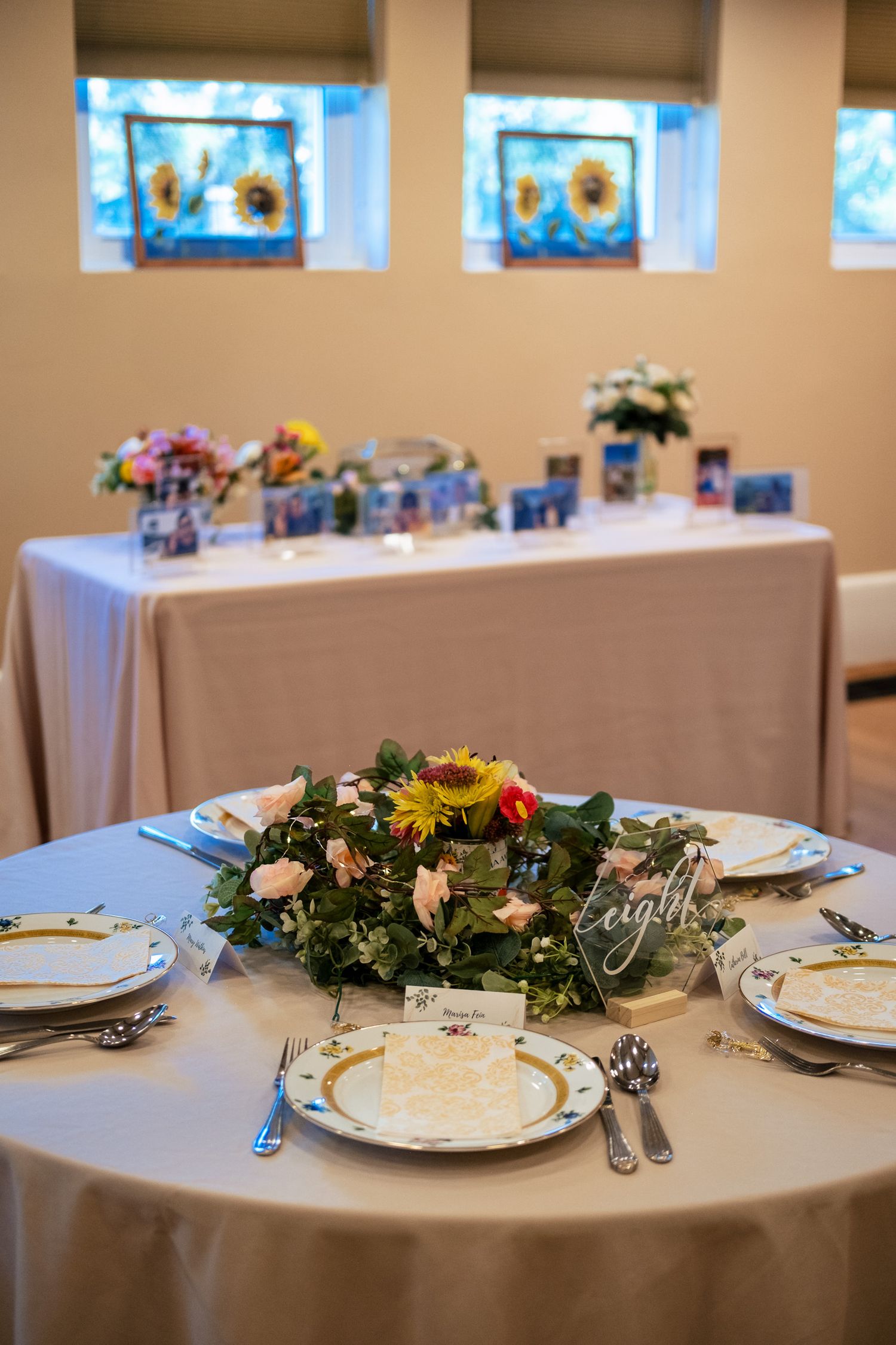 Round wedding reception table set with fine china, silverware, and a lush sunflower and pink rose centerpiece arrangement.