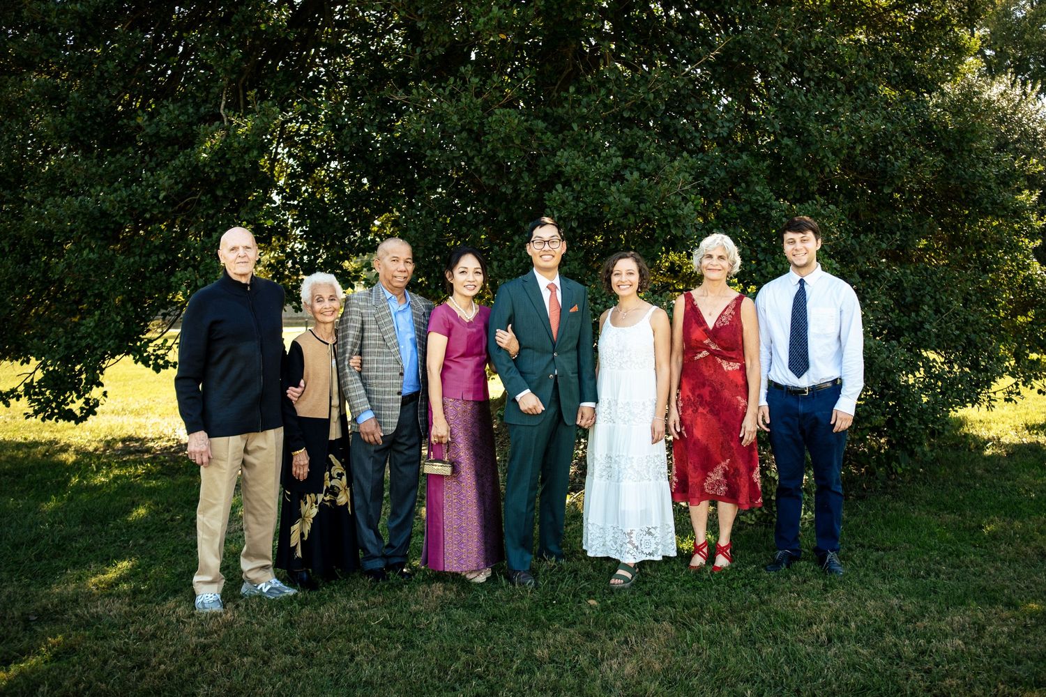 Wedding party group portrait of eight guests smiling together in front of lush green trees outdoors.
