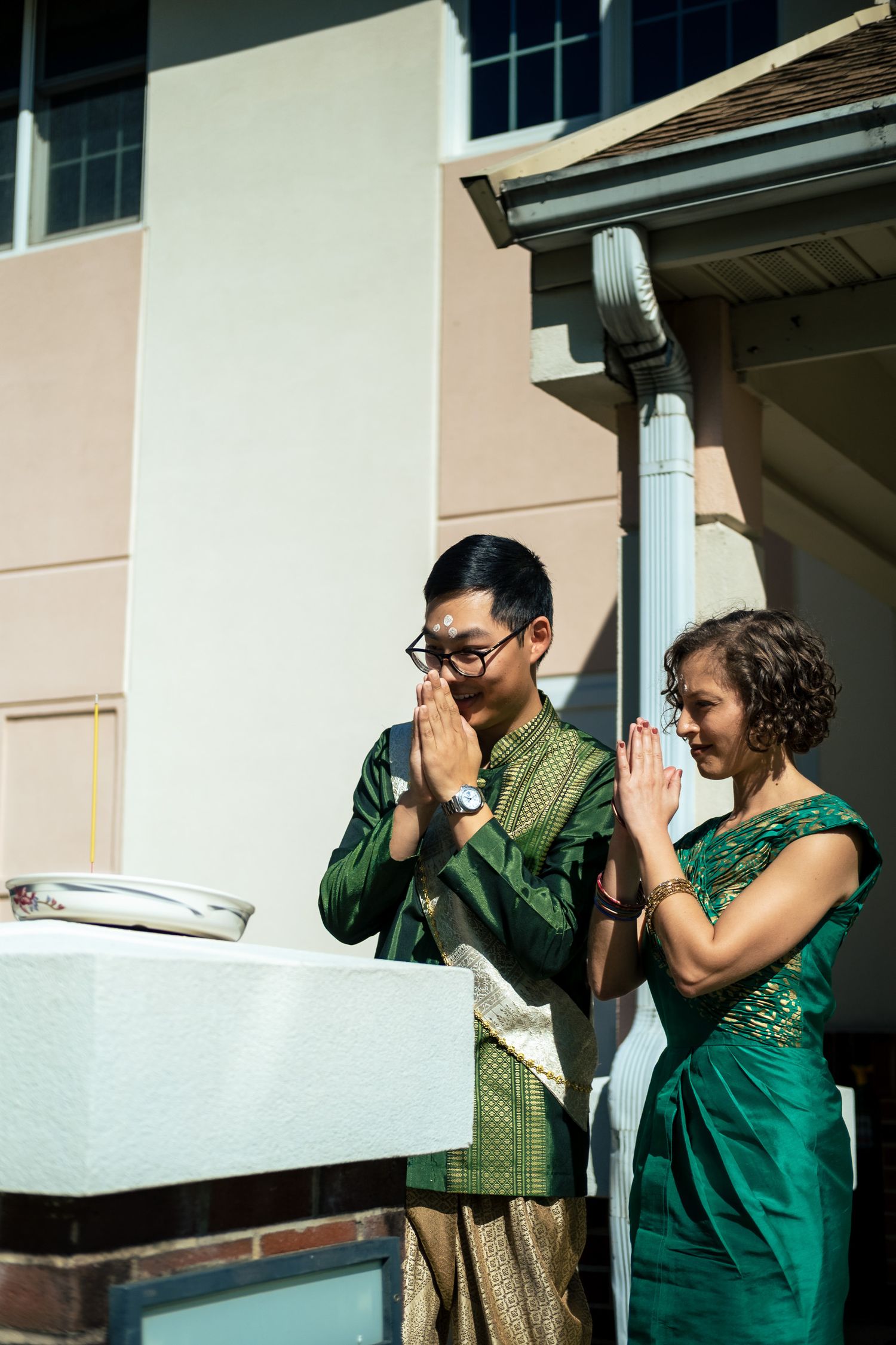 Couple in traditional Thai green wedding outfits performs respectful wai greeting gesture outdoors.