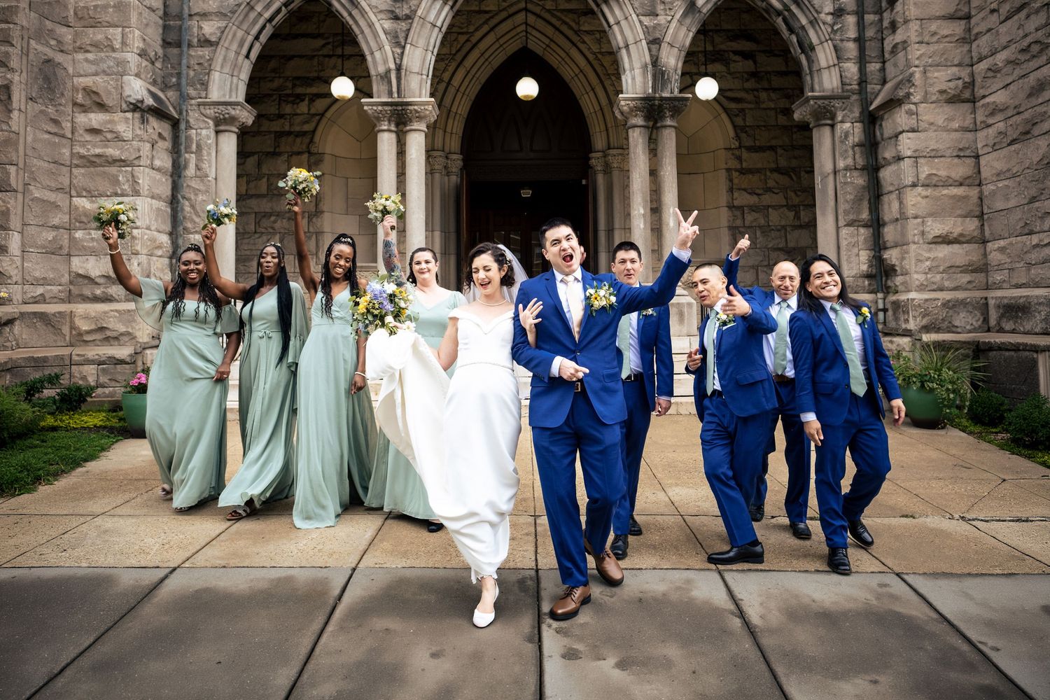 Bride, groom, and wedding party walk joyfully outside a grand stone church after the ceremony.