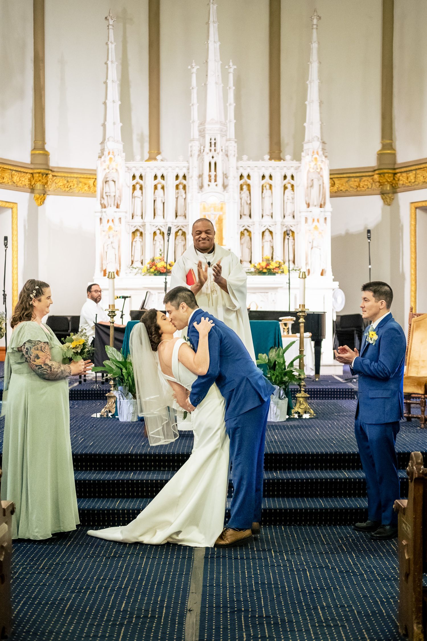 Groom dips bride for a romantic first kiss at the altar as priest and wedding party celebrate.