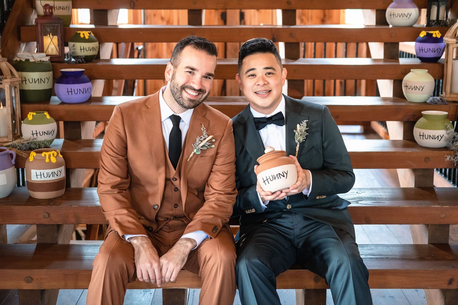 Two grooms in suits pose with a Winnie the Pooh honey pot at their Winnie the Pooh themed wedding.