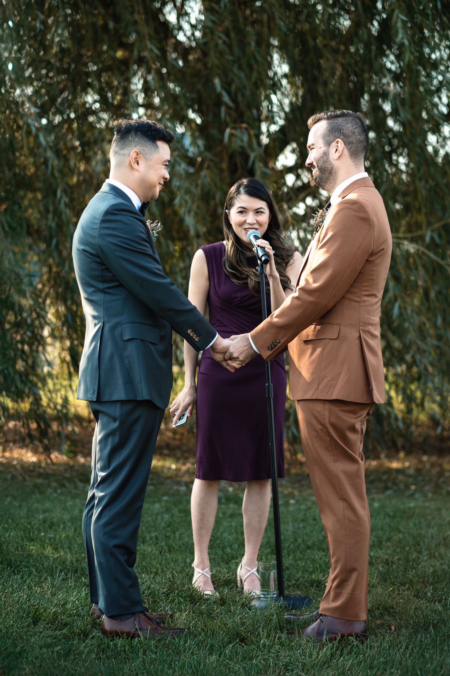 Two grooms hold hands and smile during their outdoor wedding ceremony with a smiling female officiant.