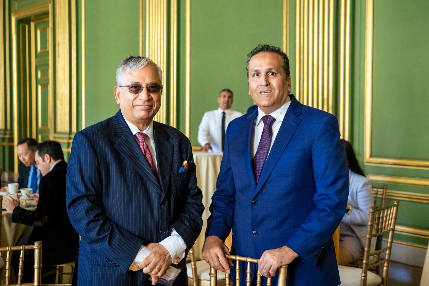 Two men in suits pose together in an ornate green and gold ballroom during a formal gathering.