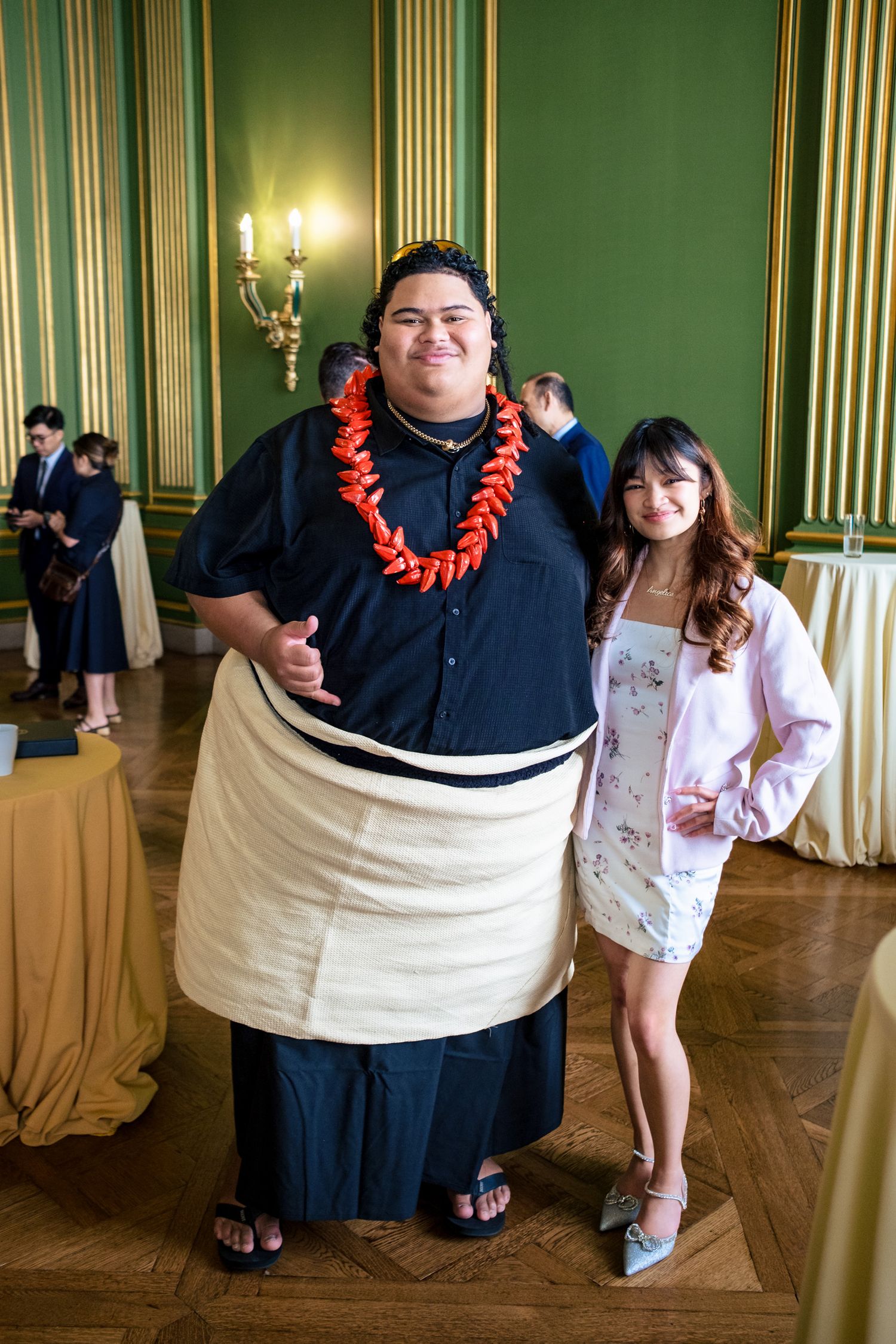 Two people posing together in an elegant green room, one wearing traditional Polynesian attire with a red lei.