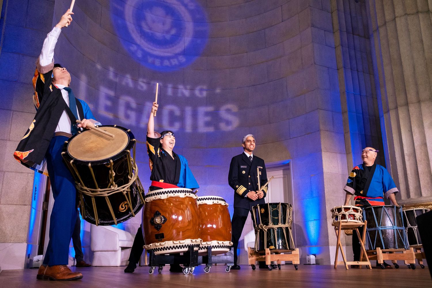 Taiko drummers perform on stage at a Lasting Legacies event, with blue lighting and a ceremonial backdrop.