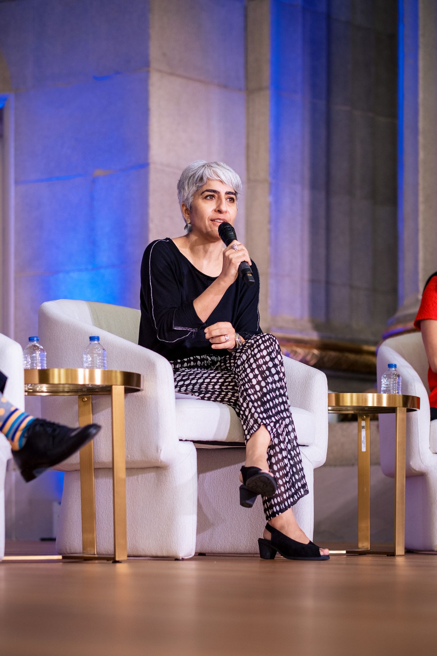 Woman with short silver hair speaks into microphone at panel discussion, seated in white chair on stage.