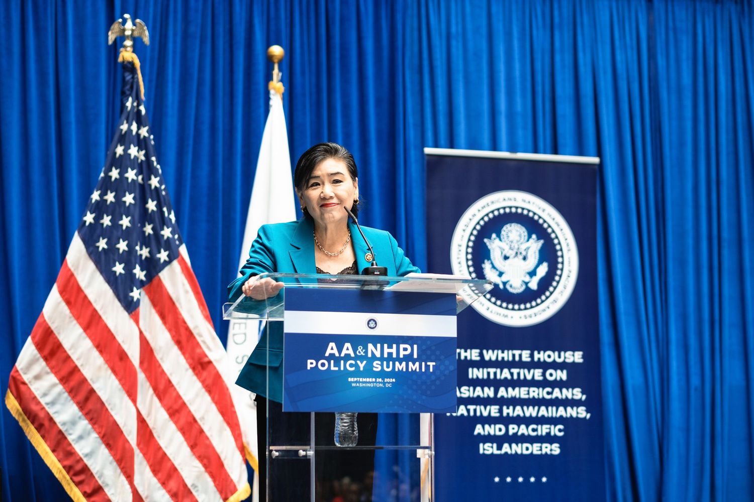 Woman in teal jacket speaks at AA&NHPI Policy Summit podium with American flag and White House Initiative banner.