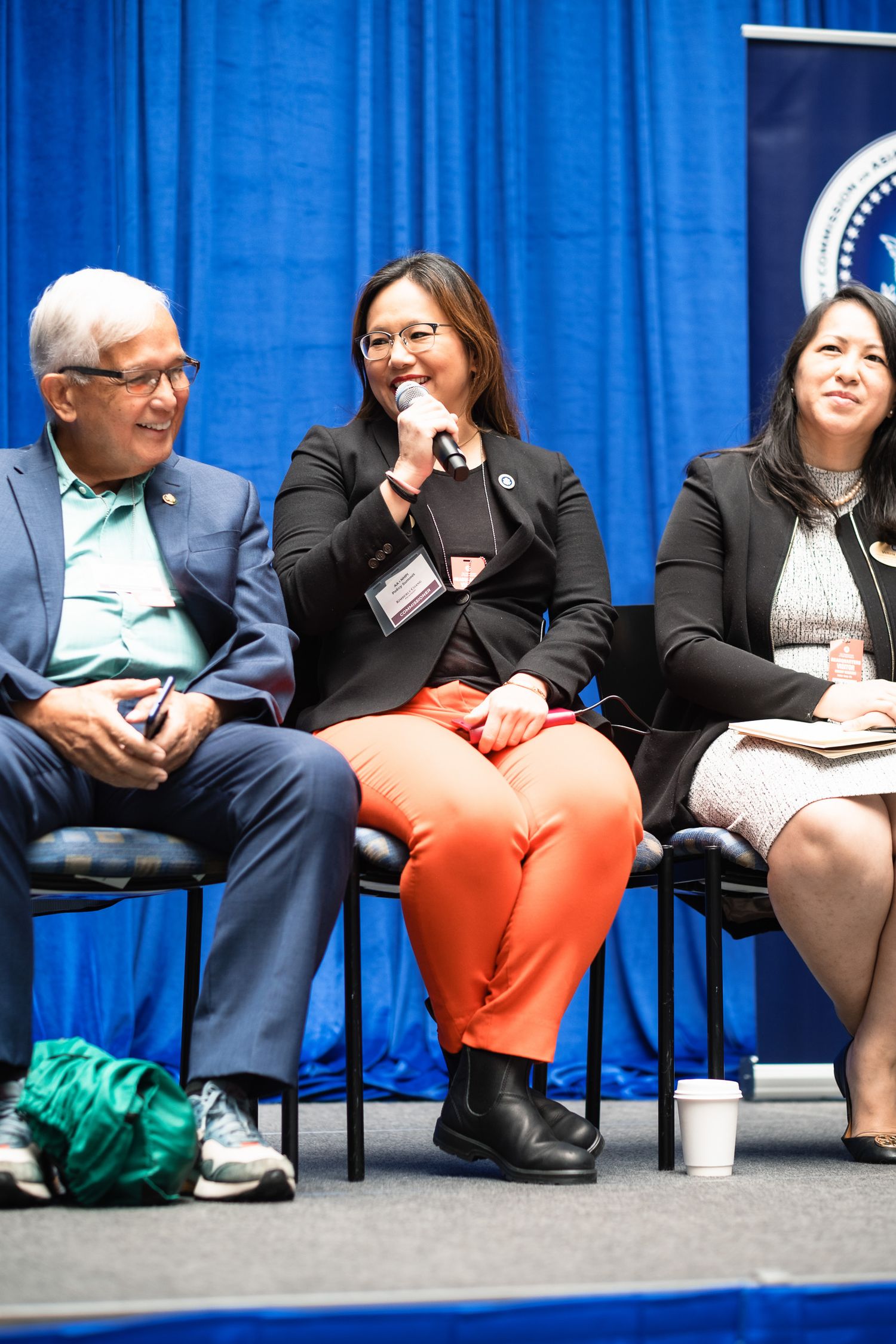 Three professionals seated on stage at a formal panel event, with blue backdrop and organizational seal visible.