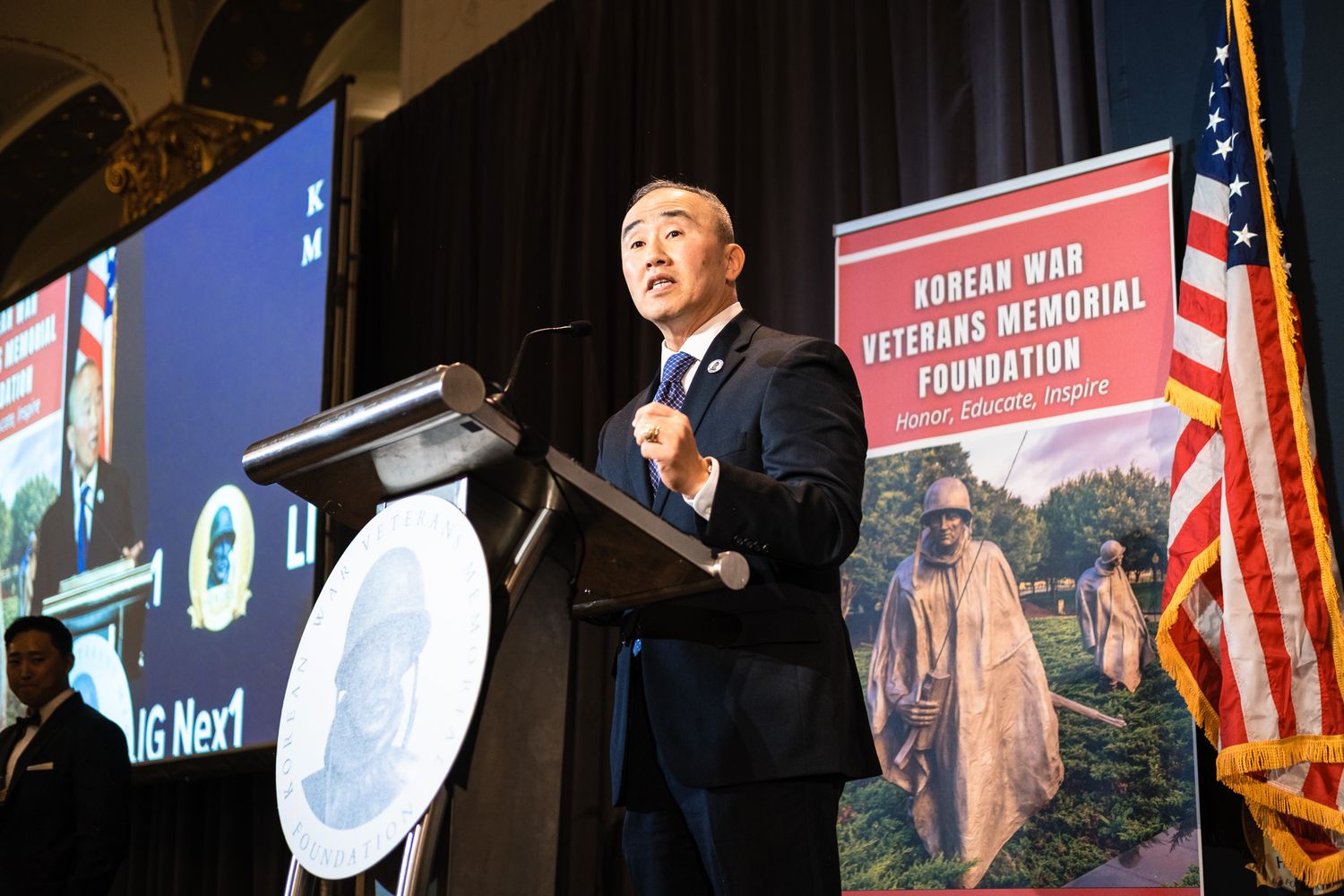 Man speaks at podium for Korean War Veterans Memorial Foundation event with US flag and memorial banner backdrop.