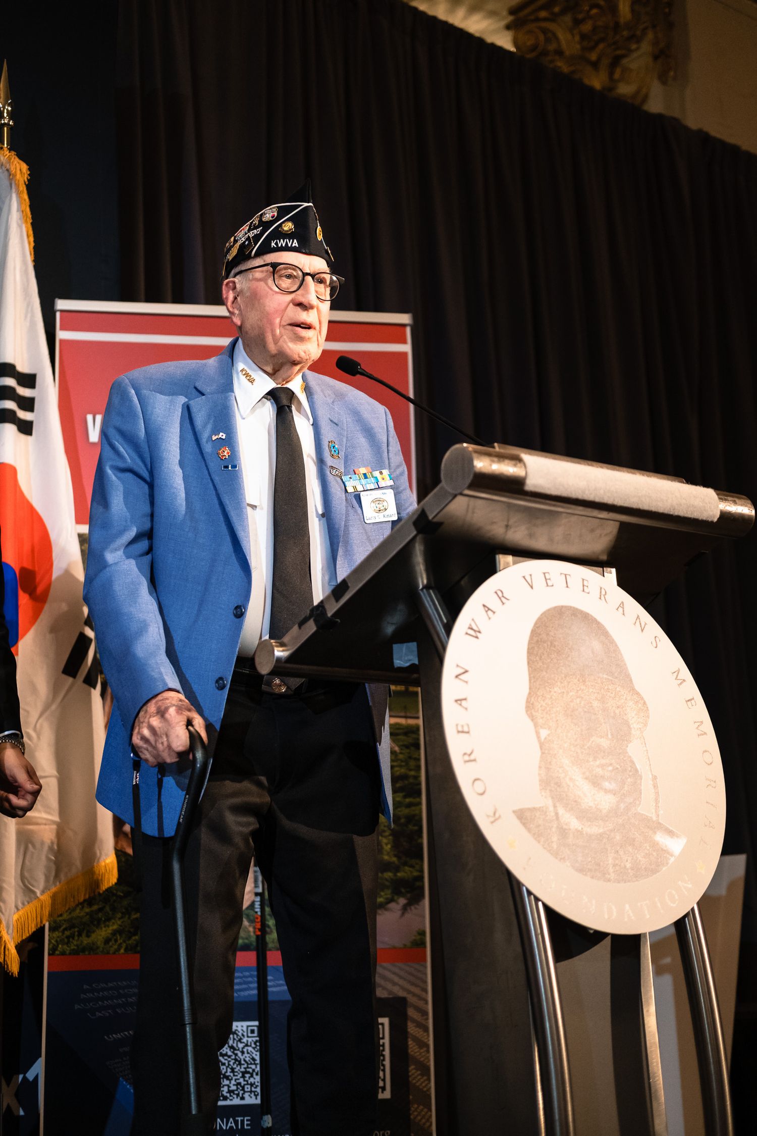 Korean War veteran speaking at podium during commemorative ceremony with South Korean flag.
