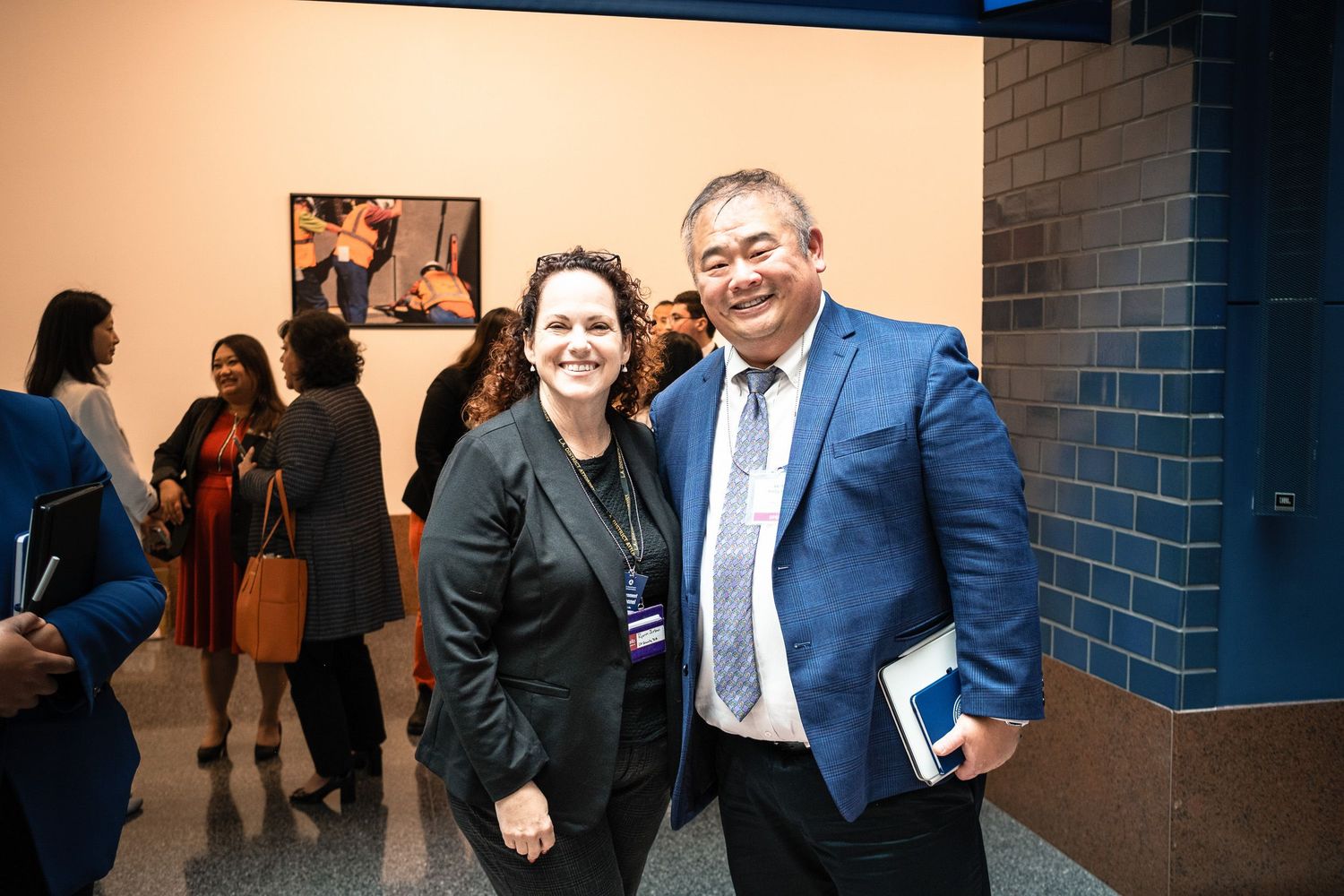 Two professionals smile at a formal networking event, man in blue suit and woman in black jacket, with guests mingling in background.