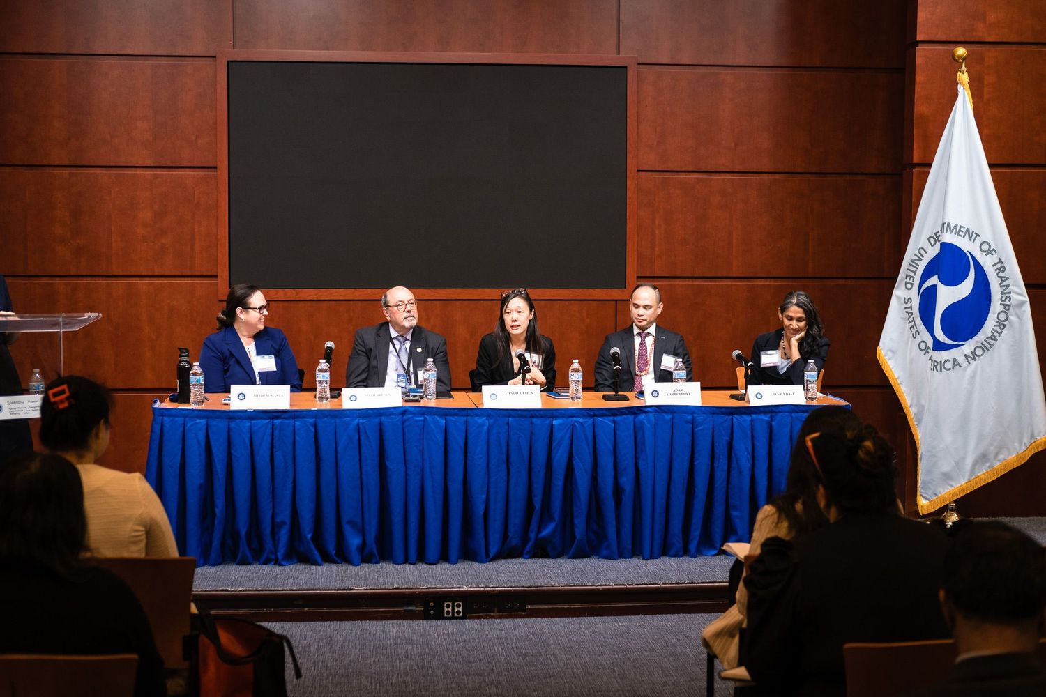 Five panelists seated at a blue-draped table during a formal conference panel discussion in a wood-paneled room.