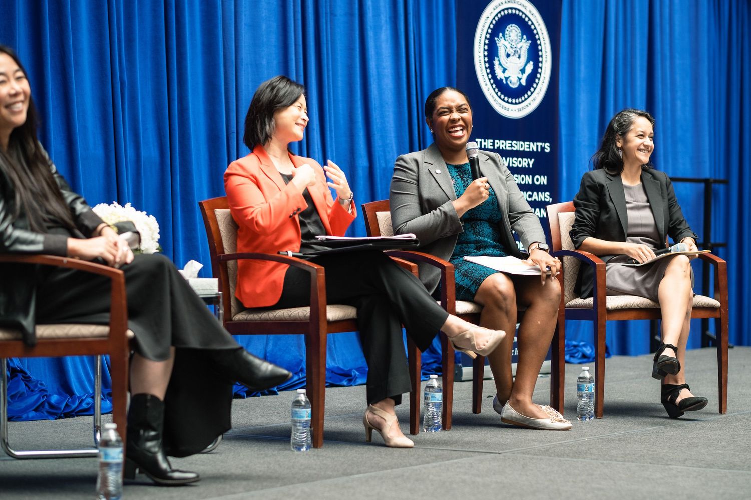 Four professional women laugh and converse during a panel discussion at a Presidential Advisory Commission event.