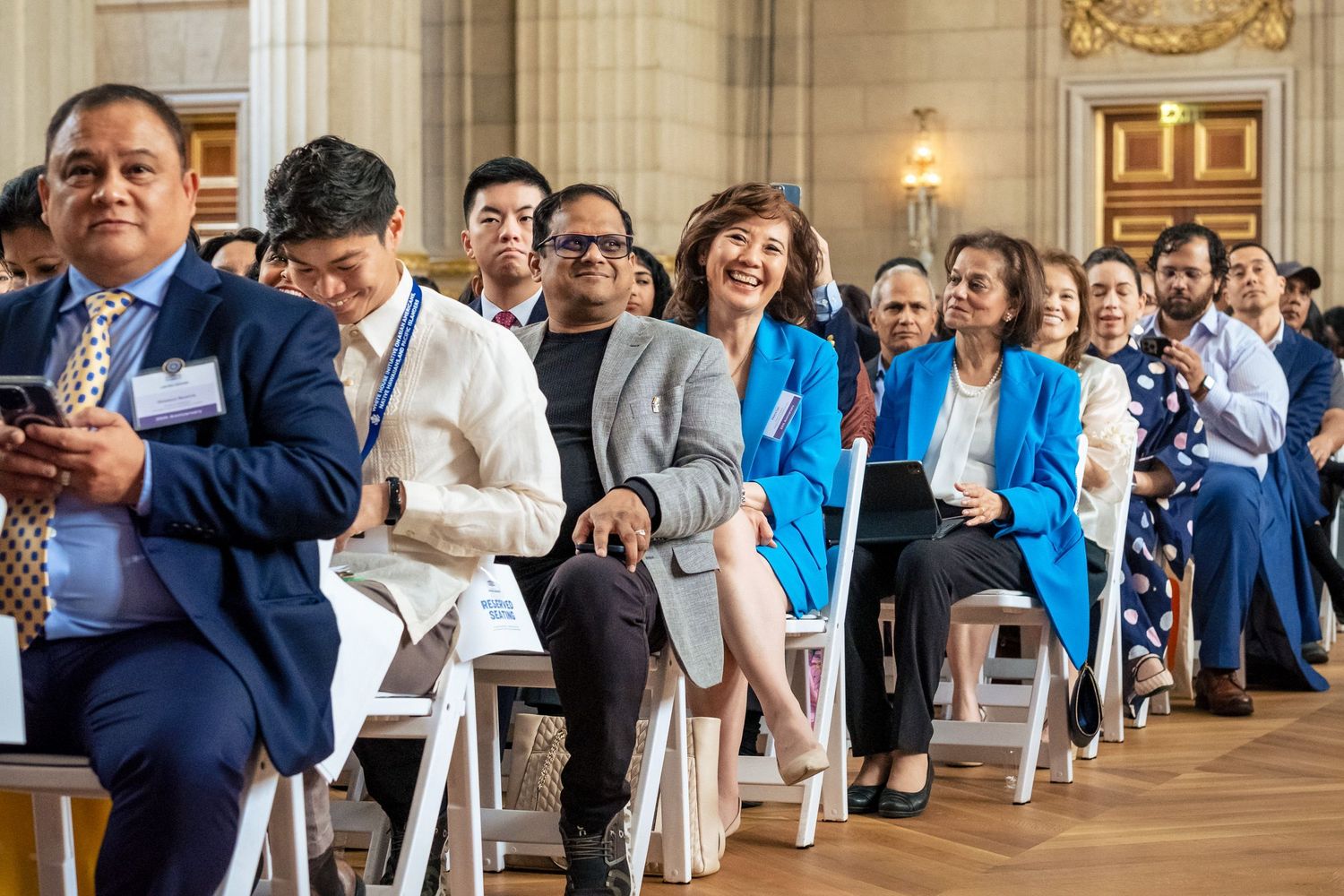 Diverse group of smiling attendees seated in rows at a formal indoor event in an elegant hall.