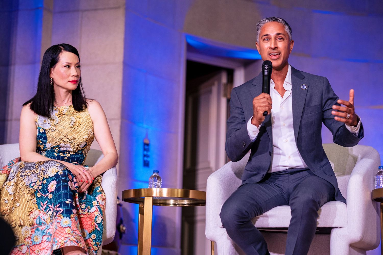Two speakers seated on stage at a formal event, woman in floral dress and man in blue suit gesturing during panel discussion.