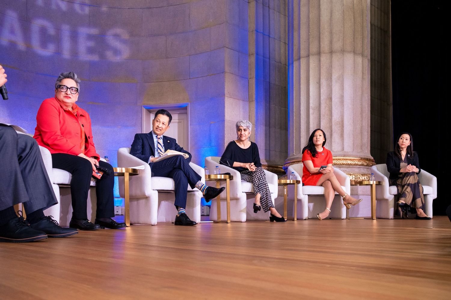Panel of six professionals seated on stage at a formal conference event with decorative backdrop.