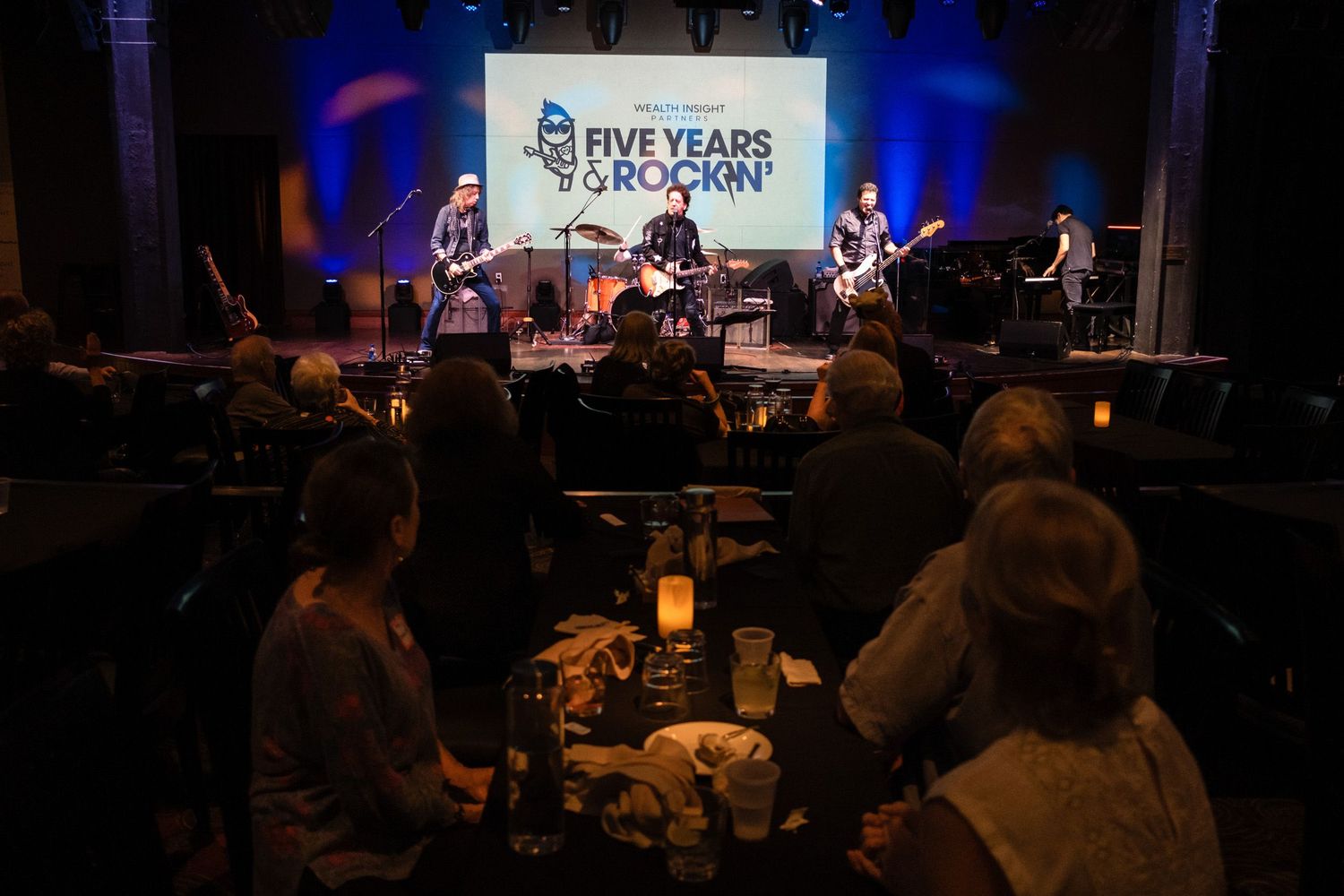 Band performs on stage at Five Years Rockin anniversary concert with audience seated at candlelit tables in dark venue.